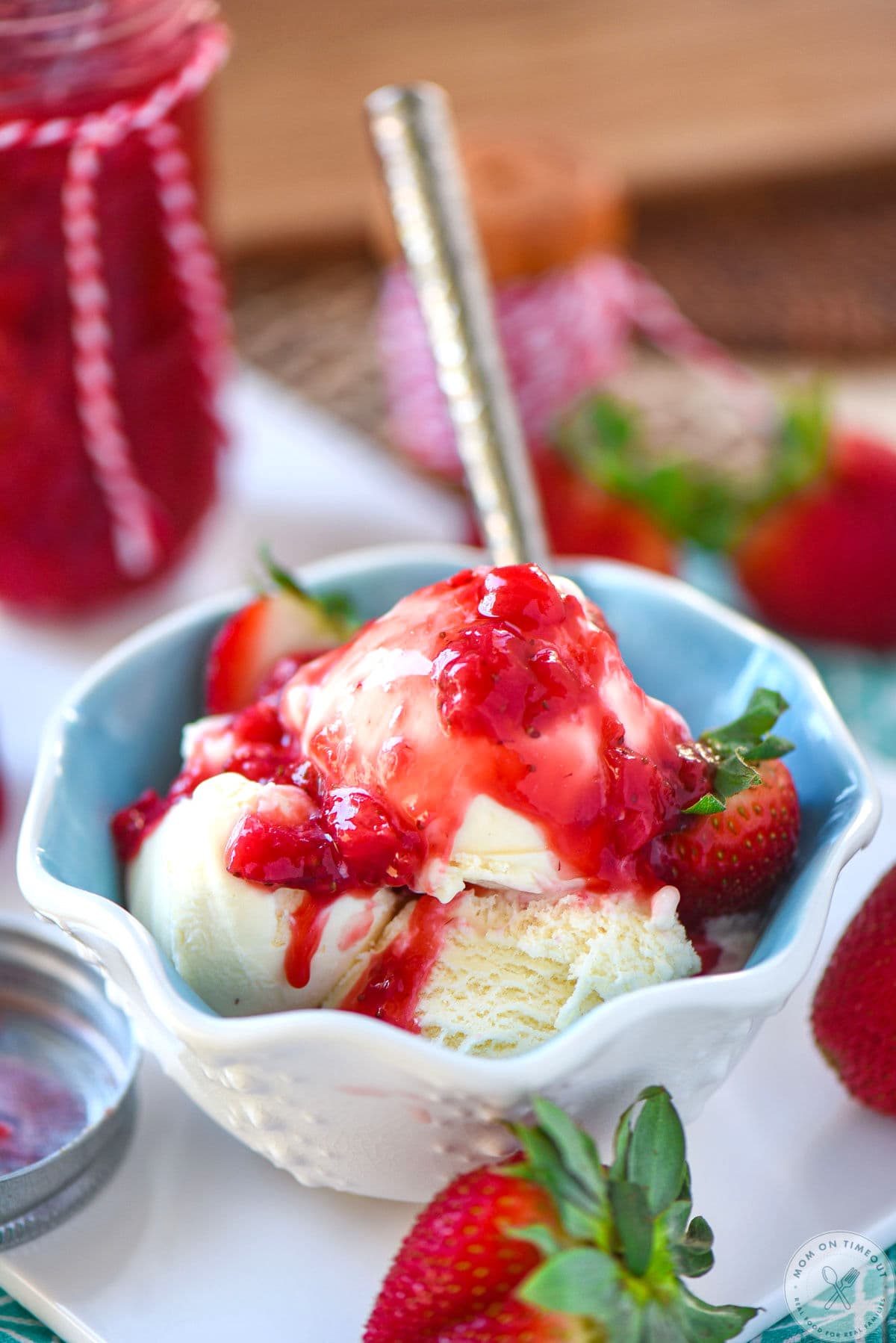 Pulled back shot of wavy ice cream bowl filled with vanilla ice cream and topped with strawberry topping for cheesecake or other desserts. Spoon is in the bowl and a jar of the strawberry sauce can be seen in the background.