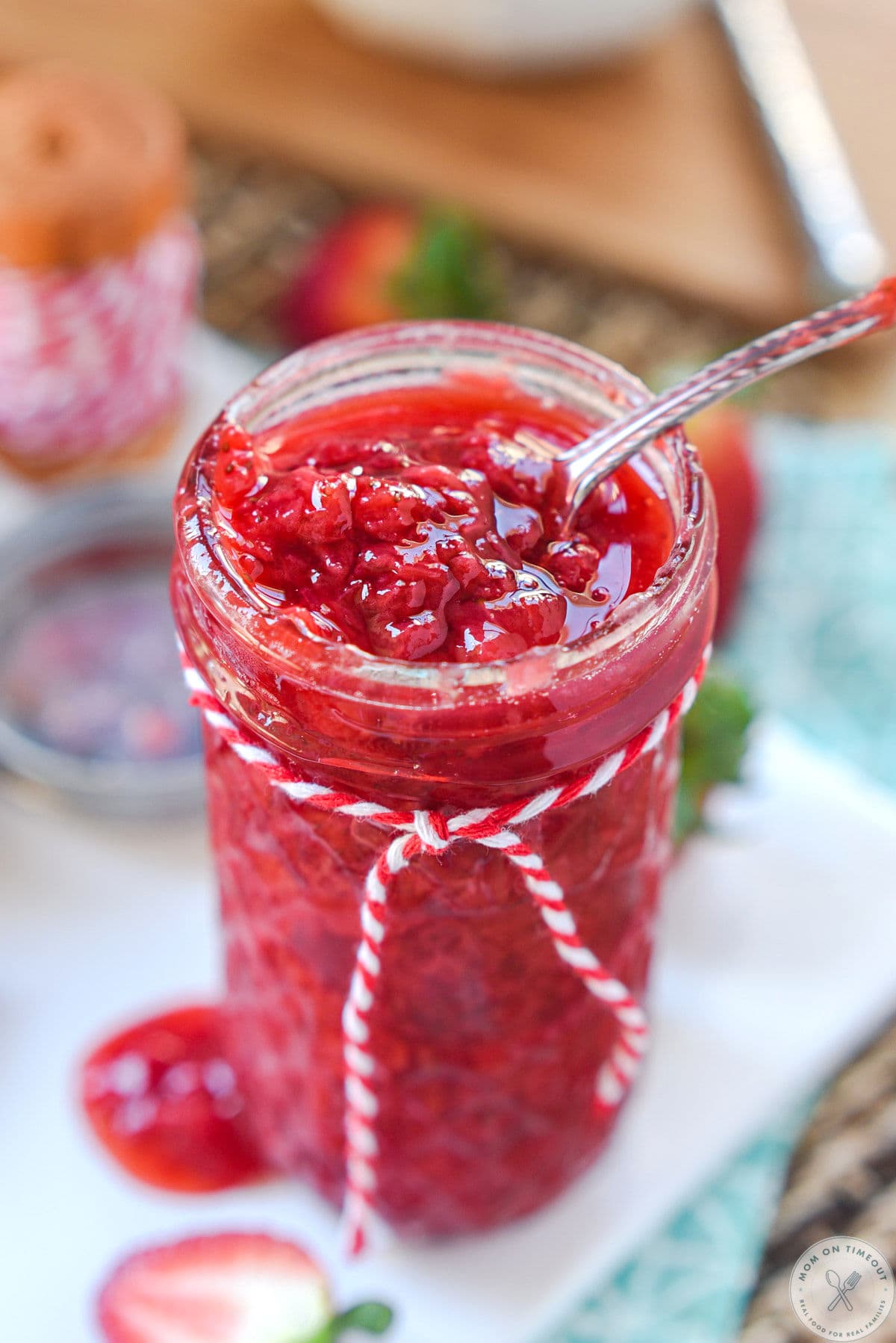 Glass canning jar filled with strawberry topping and a spoon is resting in the sauce. Jar is tied with red and white striped twine.