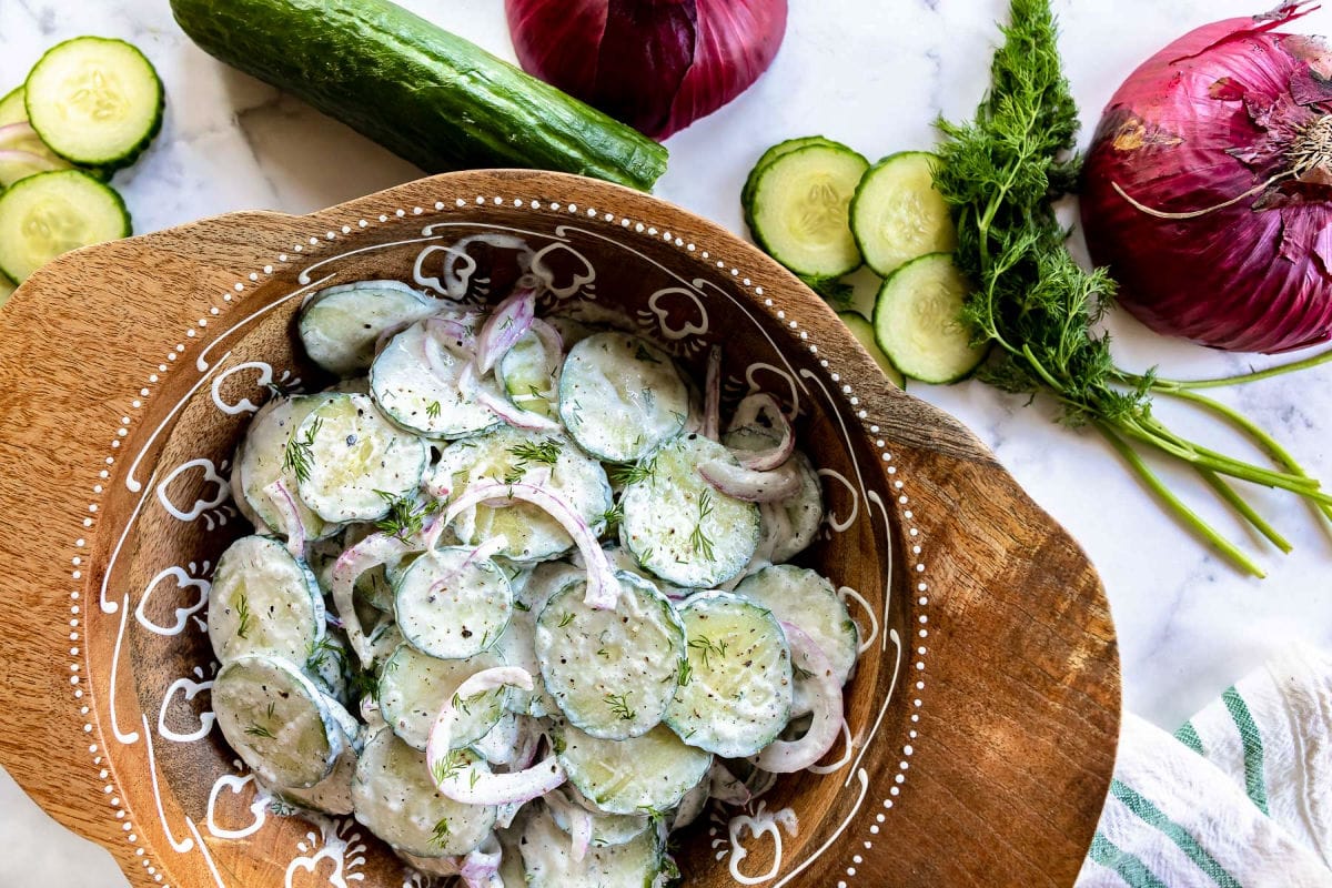 top down view of cucumber salad in wooden bowl with cut cucumber dill and red onion on marble surface