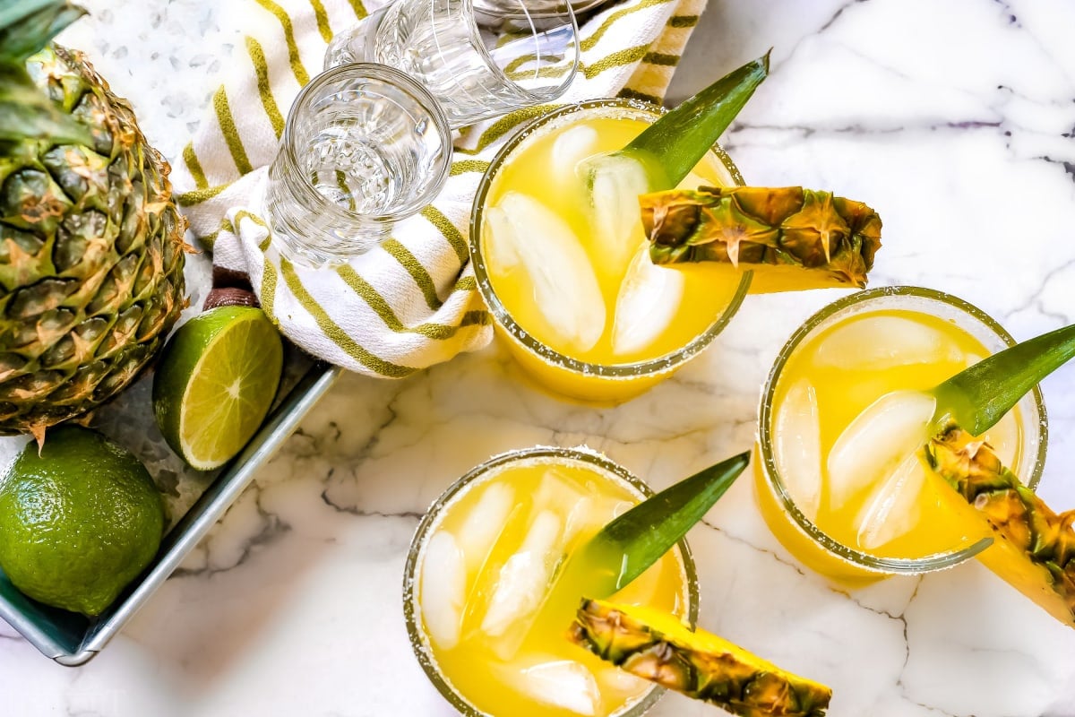 easy pineapple margarita recipe in glasses next to whole pineapple and limes on white marble backdrop