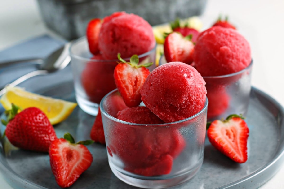 easy strawberry sorbet recipe shown in three small glasses on gray plate with halved strawberries as garnish