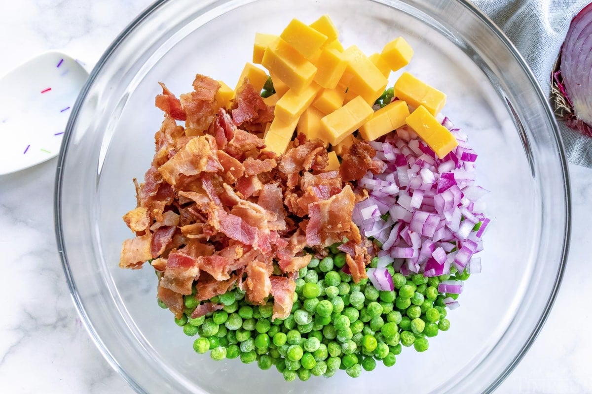 pea salad ingredients in a clear mixing bowl including peas red onions cheese and bacon
