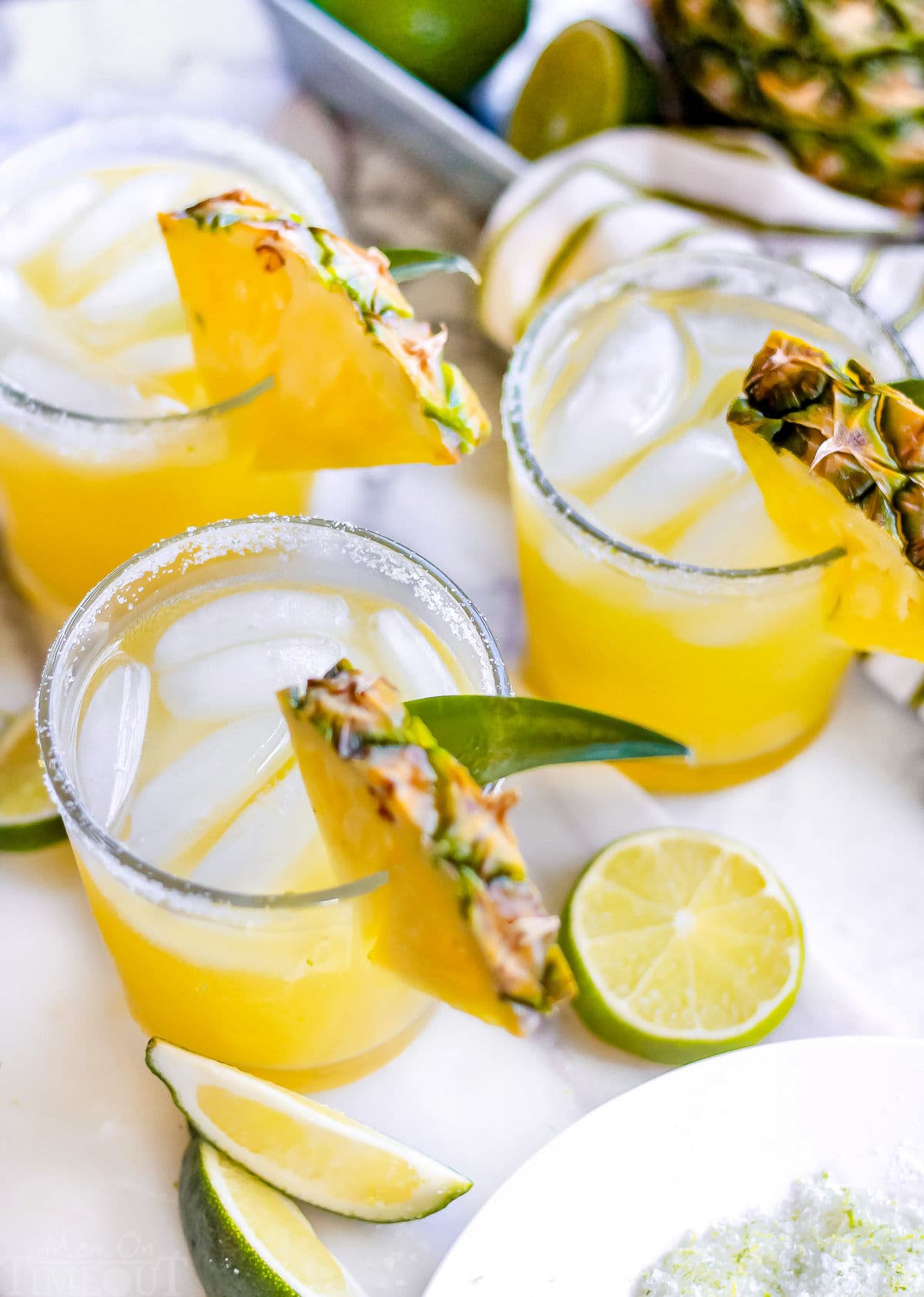 pineapple margaritas on marble surface with lime and pineapple wedges next to bowl of lime margarita salt