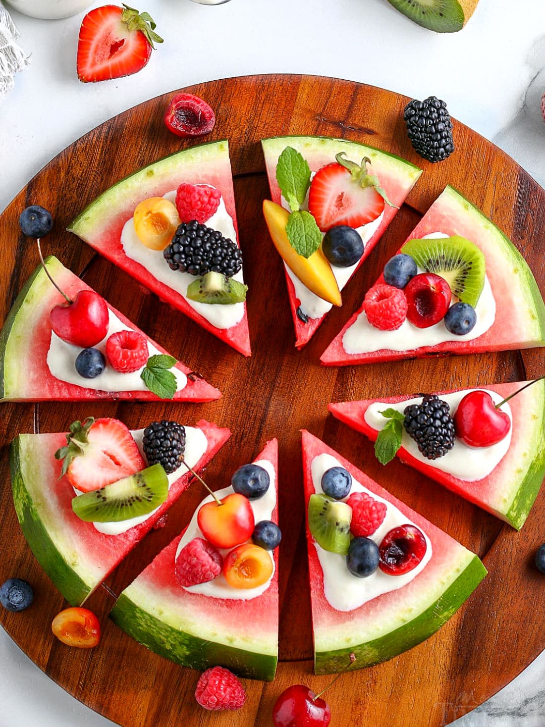 whole watermlon pizza cut into wedges topped with cream cheese glaze and fresh fruit on wood board