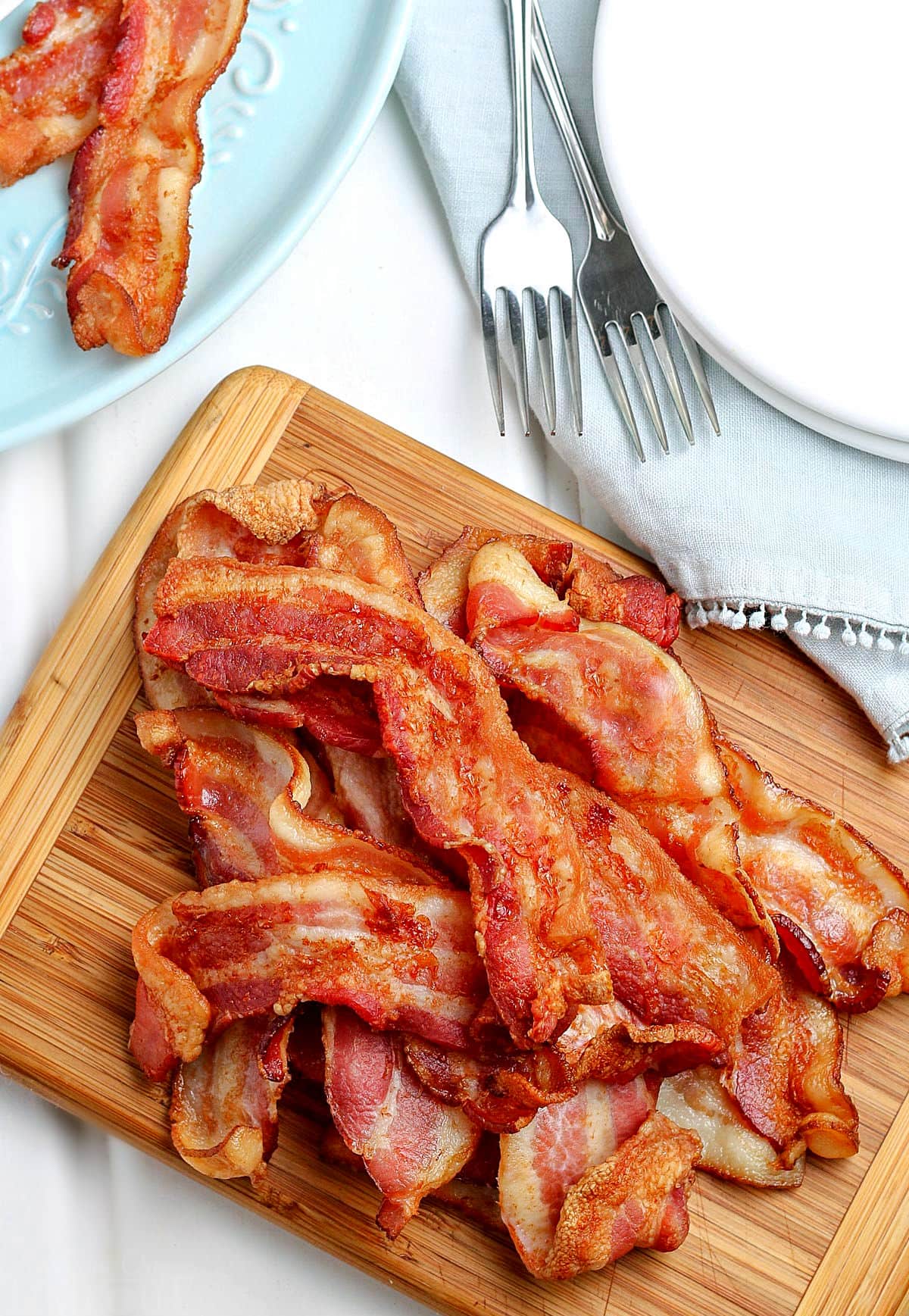 top down view of air fryer bacon piled up on wood cutting board with stack of white plates to the side