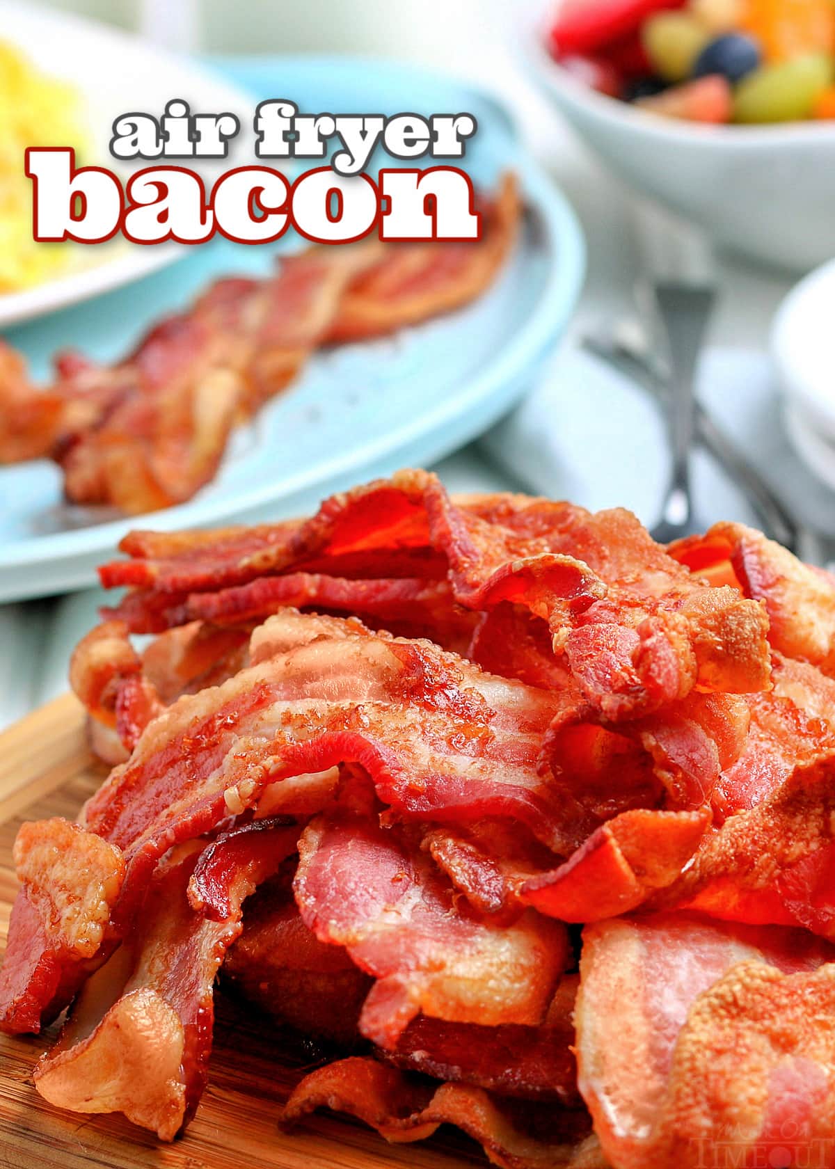 air fryer bacon piled high on wood board with blue blue plate in background with eggs and bacon on it and bowl of fruit and text overlay up top