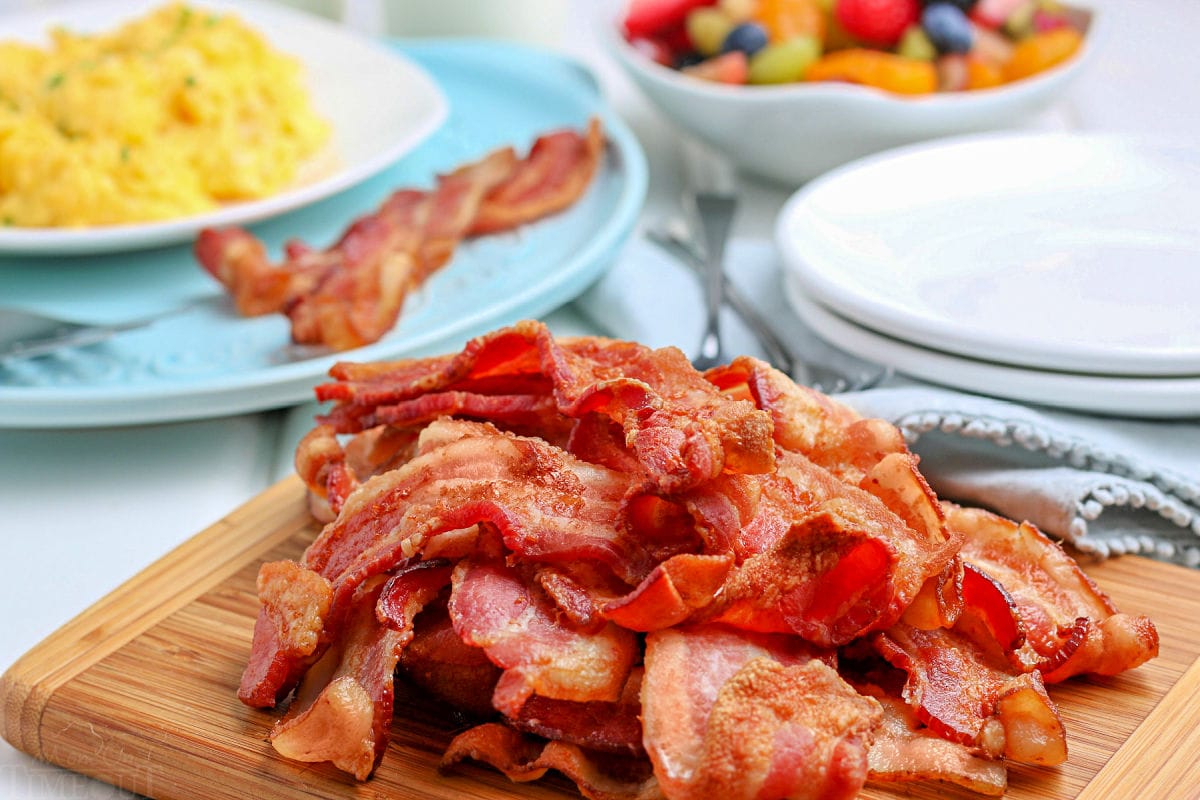 bacon piled high on wood cutting board with bowl of fruit salad and plate with eggs and bacon in the background