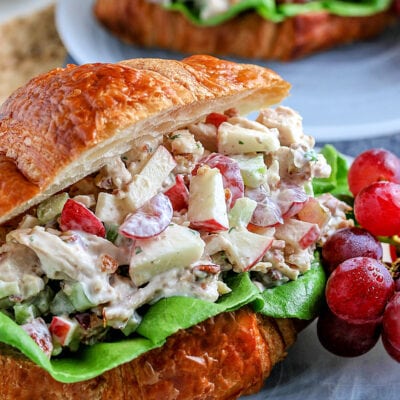 chicken salad on croissant sitting on glass plate with grapes on the side blue napkin underneath squared