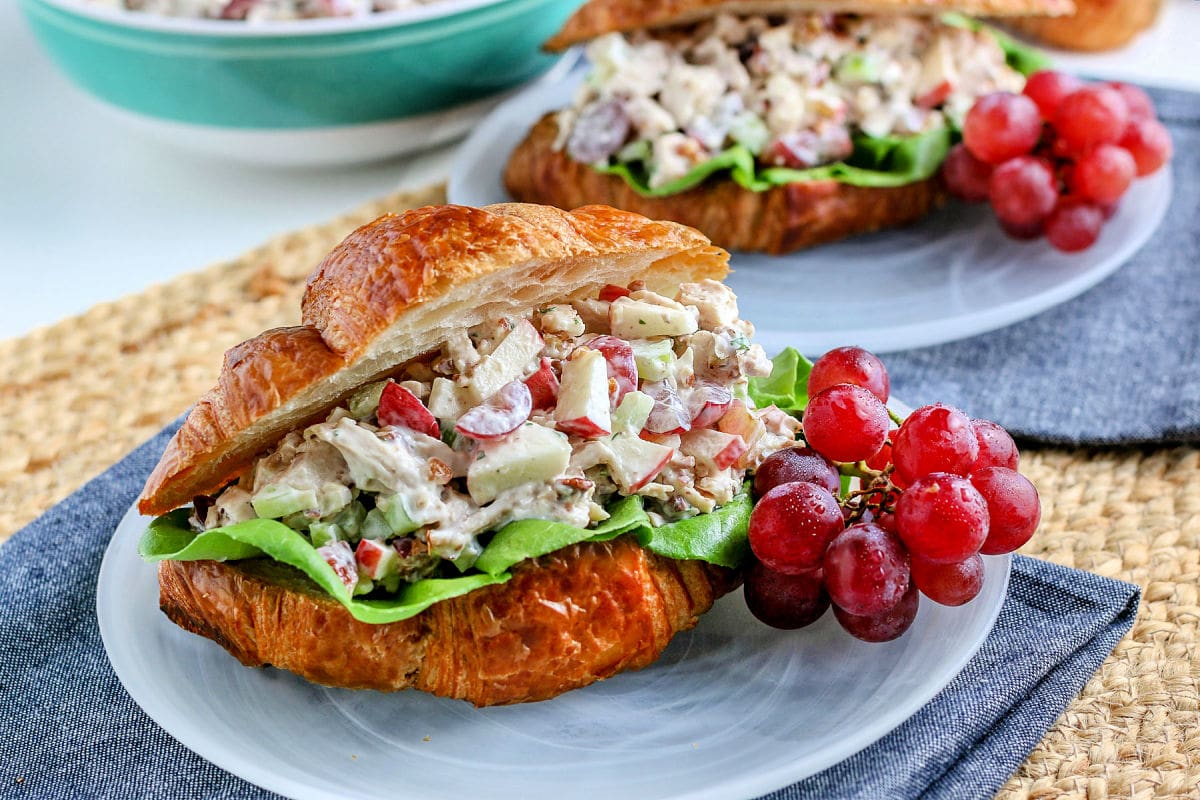 chicken salad on croissant sitting on glass plate with grapes on the side blue napkin underneath