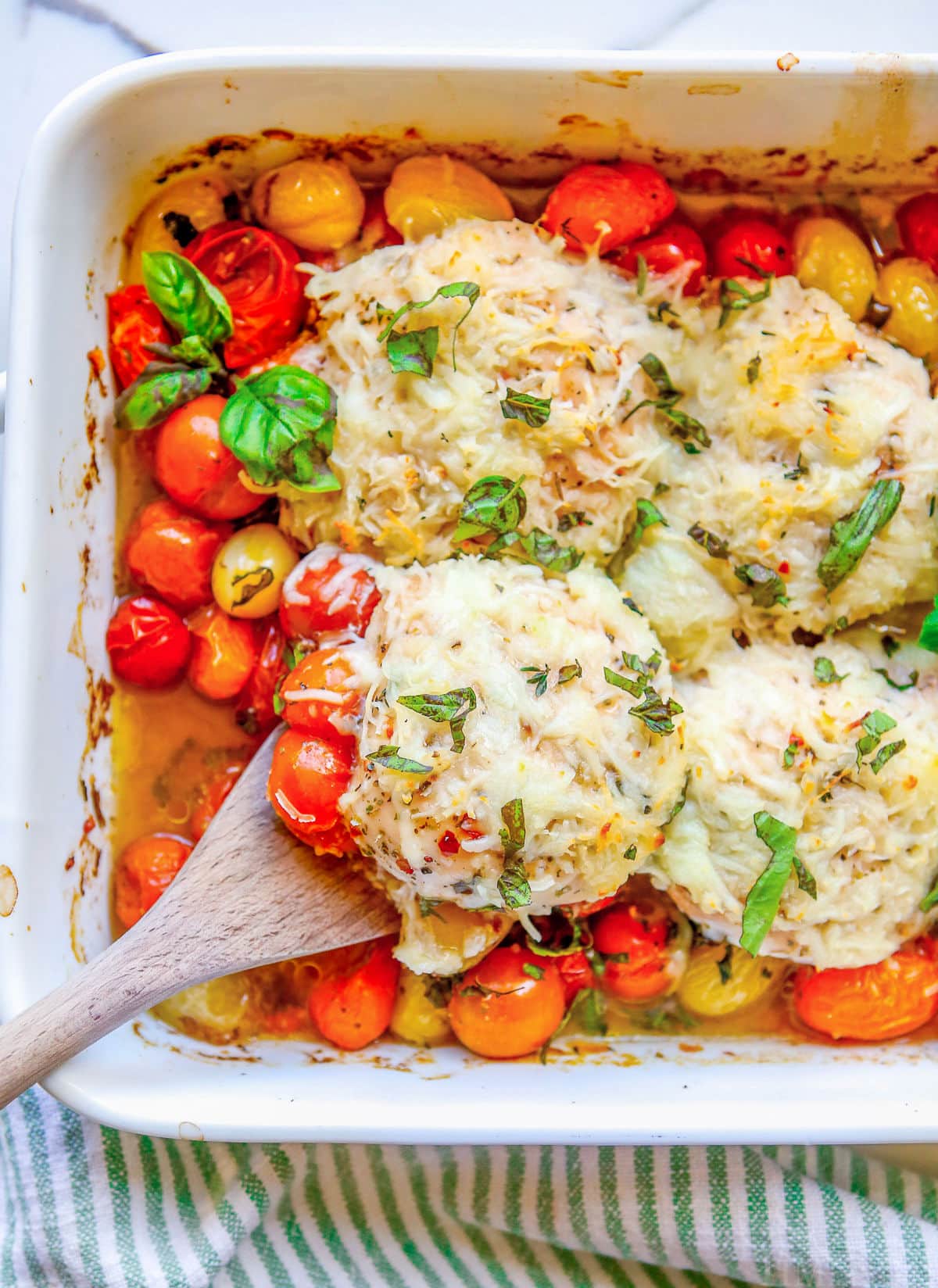top down view of baked chicken breasts in white casserole dish with tomatoes basil and cheese and wooden serving spoon