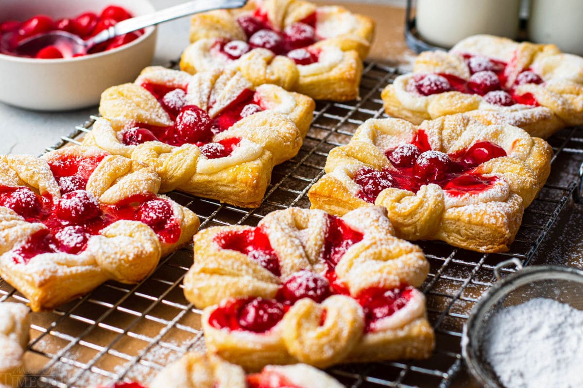 6 danishes with cherries baked and sprinkled with powdered sugar sitting on wire cooling rack