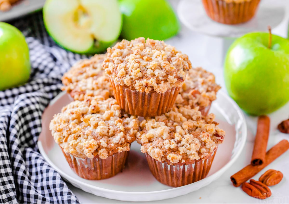 apple muffins stacked on white plate with blue and white napkin beneath the plate apples in the background