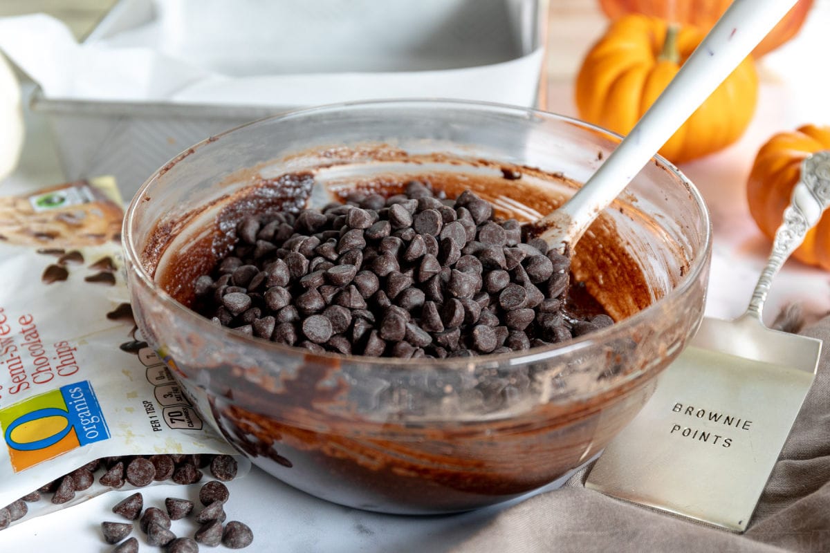 chocolate chips on top of brownie batter in glass bowl with chocolate chips scattered about and baking form in background