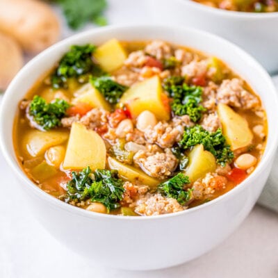 sausage kale soup in white bowl sitting on white background with spoon to the side and another bowl and potatoes in the back