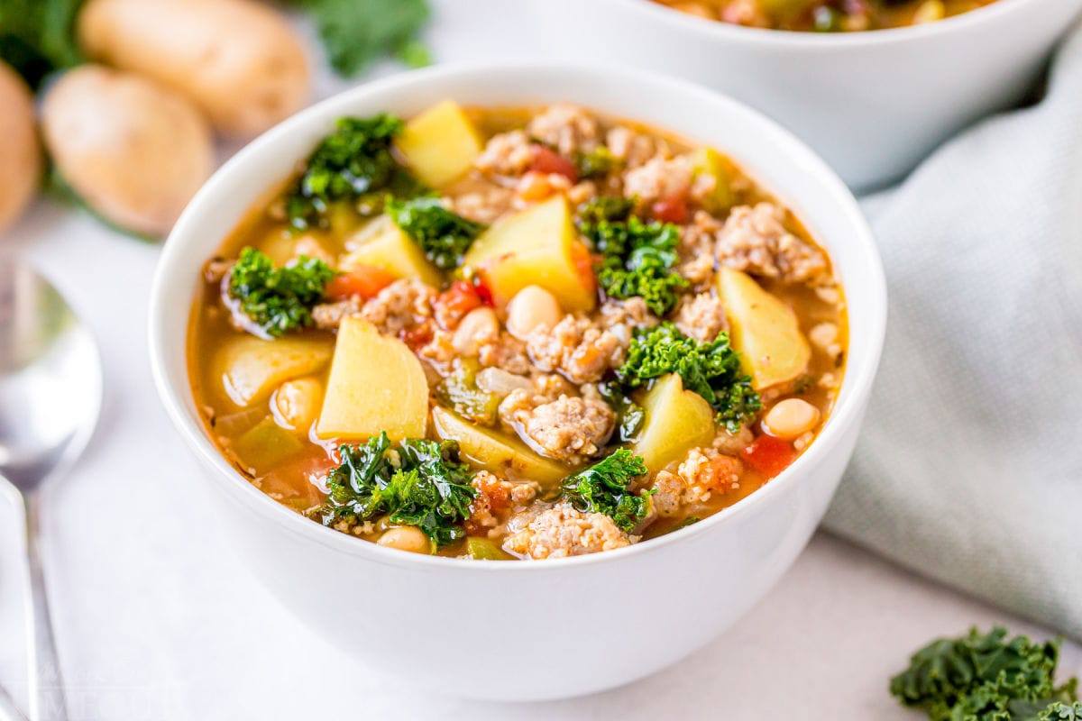 sausage kale soup in white bowl sitting on white background with spoon to the side and another bowl and potatoes in the back