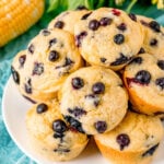 top down view of corn muffin recipe with blueberries on white plate with corn and yellow flowers behind the plate