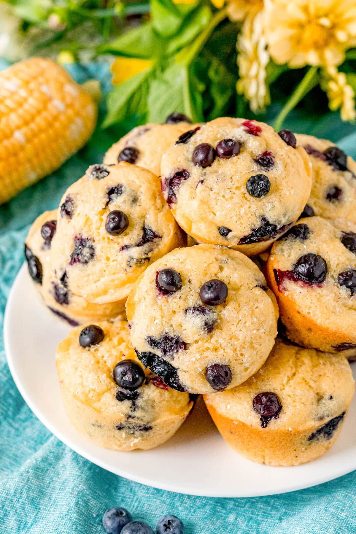 top down view of corn muffin recipe with blueberries on white plate with corn and yellow flowers behind the plate