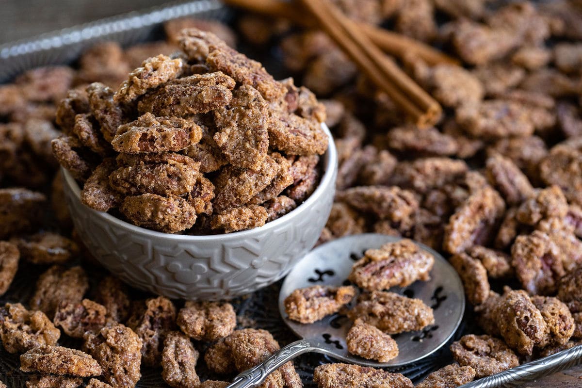 candied pecans in small bowl sitting on antique baking sheet with spoon and lots of candied pecans strewn about