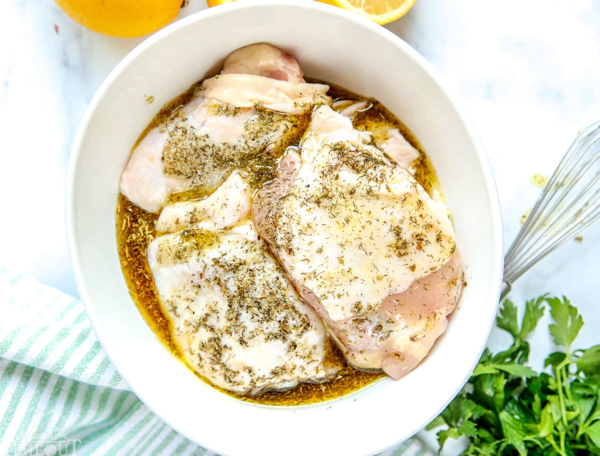 greek chicken recipe marinade poured over chicken thighs in white bowl with green and white striped towel to the side and lemons and parsley beside bowl