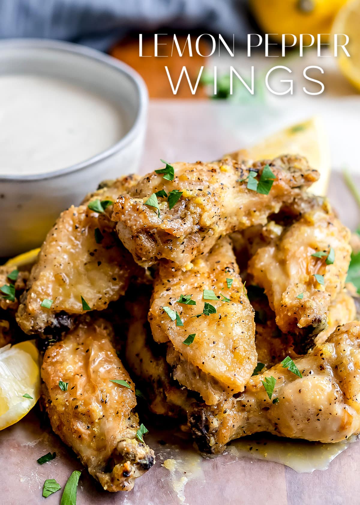 lemon pepper wings stacked in a pile with a bowl of ranch behind them and title overlay at the top