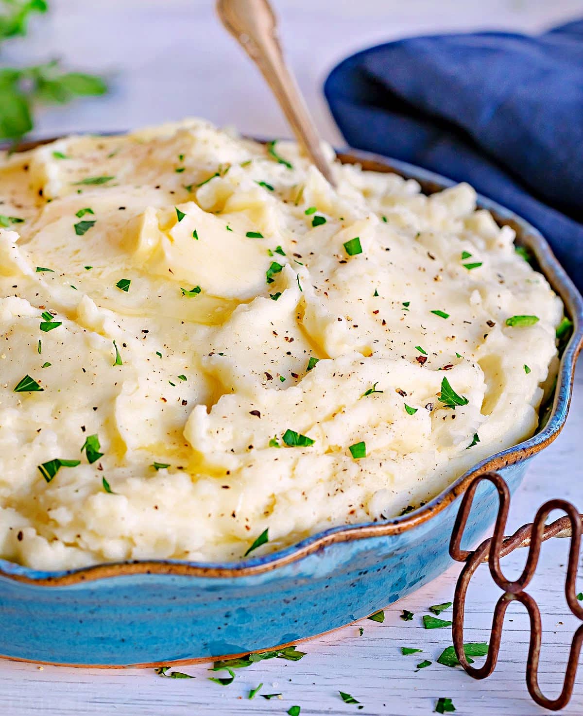 best mashed potatoes in serving bowl topped with thick slices of butter and freshly chopped parsley. blue napkin in background.