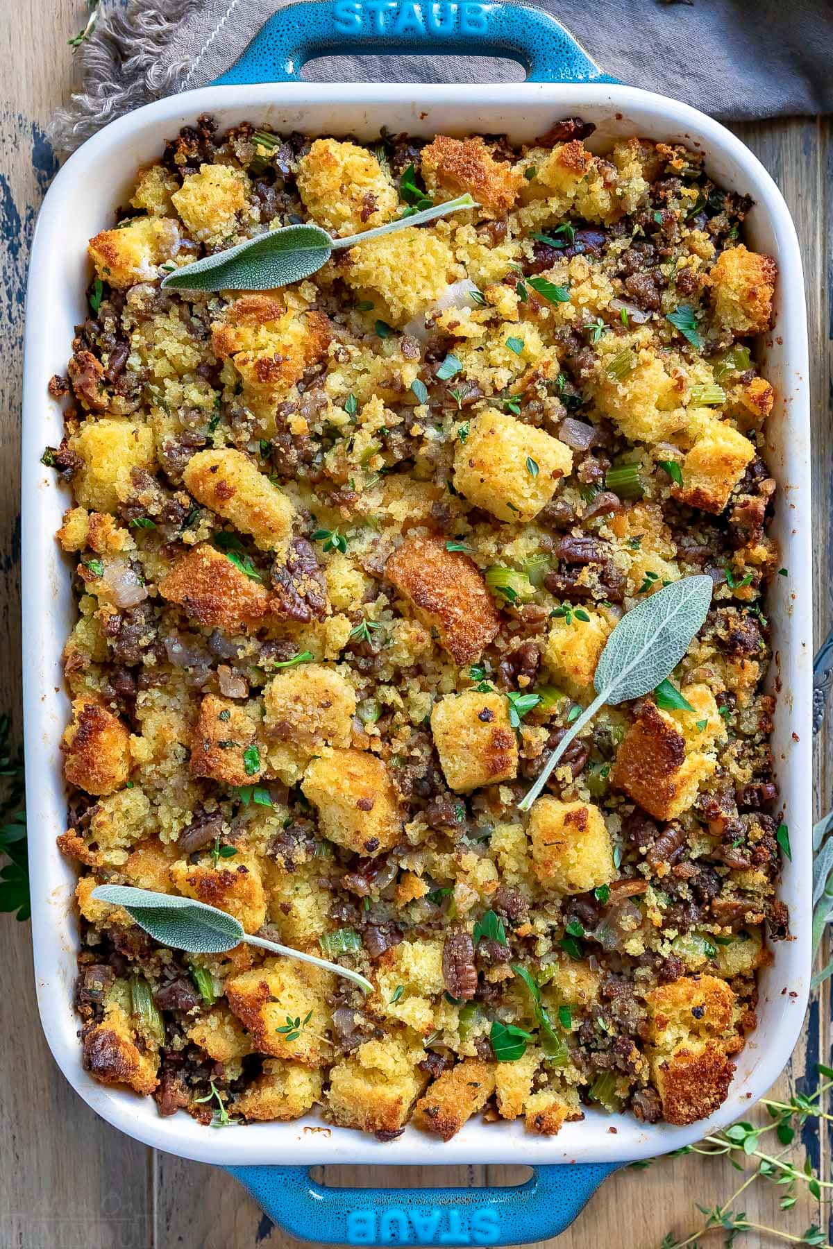 top down view of cornbread stuffing in baking dish with fresh sage leaves and thyme scattered on top