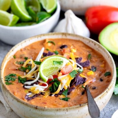 chicken enchilada soup in pottery bowl with spoon and garnished with cilantro lime tomatoes cheese and title overlay at top