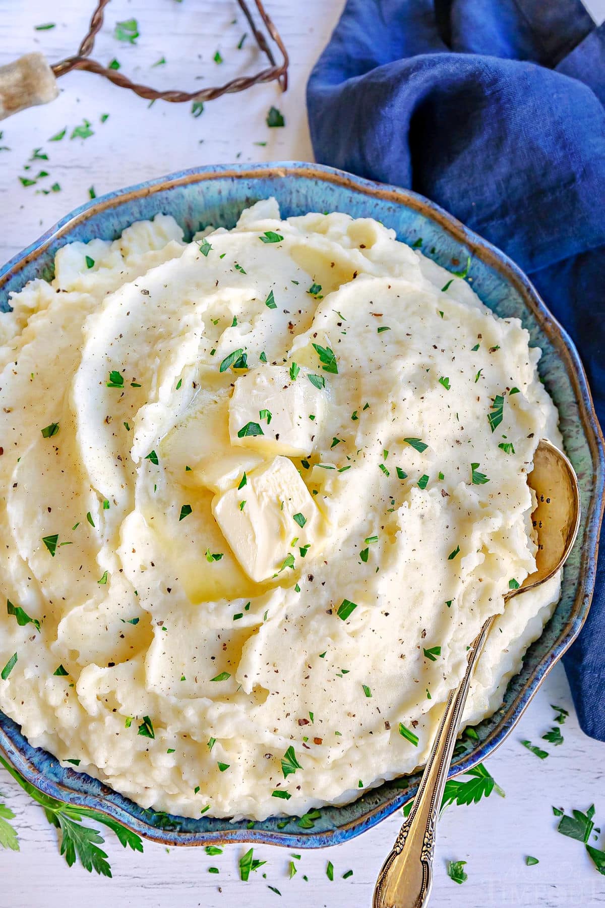 top down look at a blue and green bowl filled with fluffy mashed potatoes. slices of butter, pepper and chopped parsley garnish the mashed potatoes.