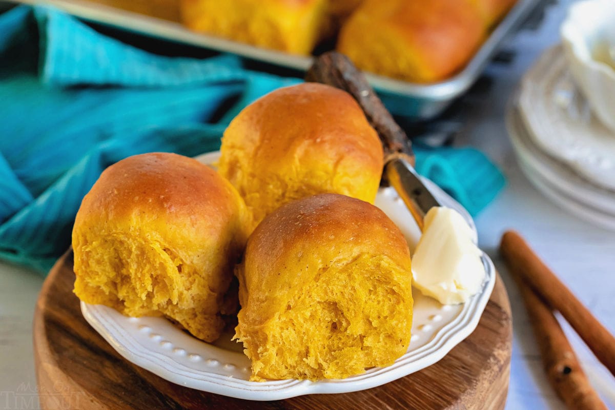 three pumpkin dinner rolls on a white plate with butter on the side on white plate with teal napkin