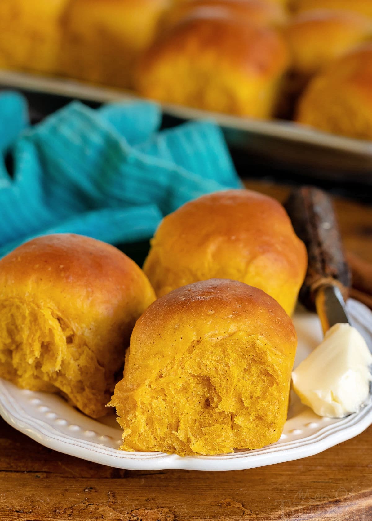 three pumpkin dinner rolls on a white plate with butter on the side and baking sheet in background