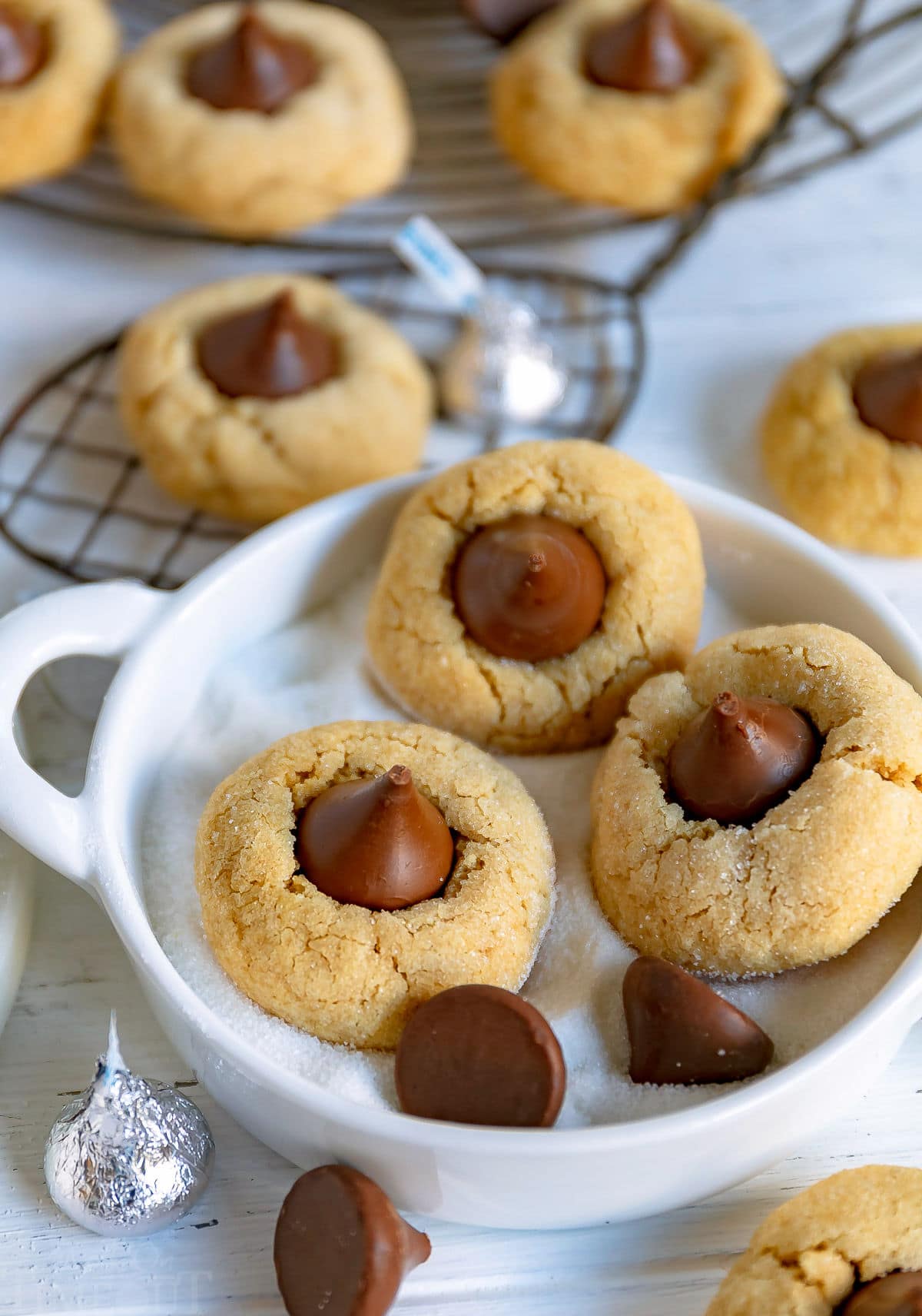 three peanut butter blossom cookies in small white bowl with sugar in it and there are more cookies in background