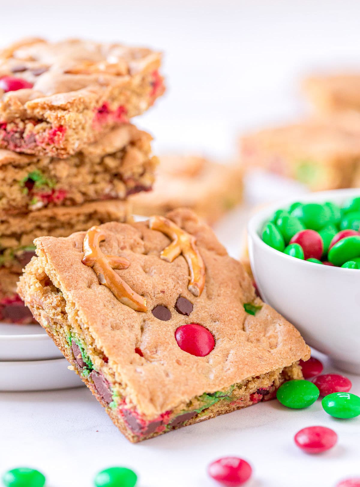 cookie bar leaned up against white plate with more cookie bars stacked on it and sitting next to a white bowl filled with red and green M&Ms