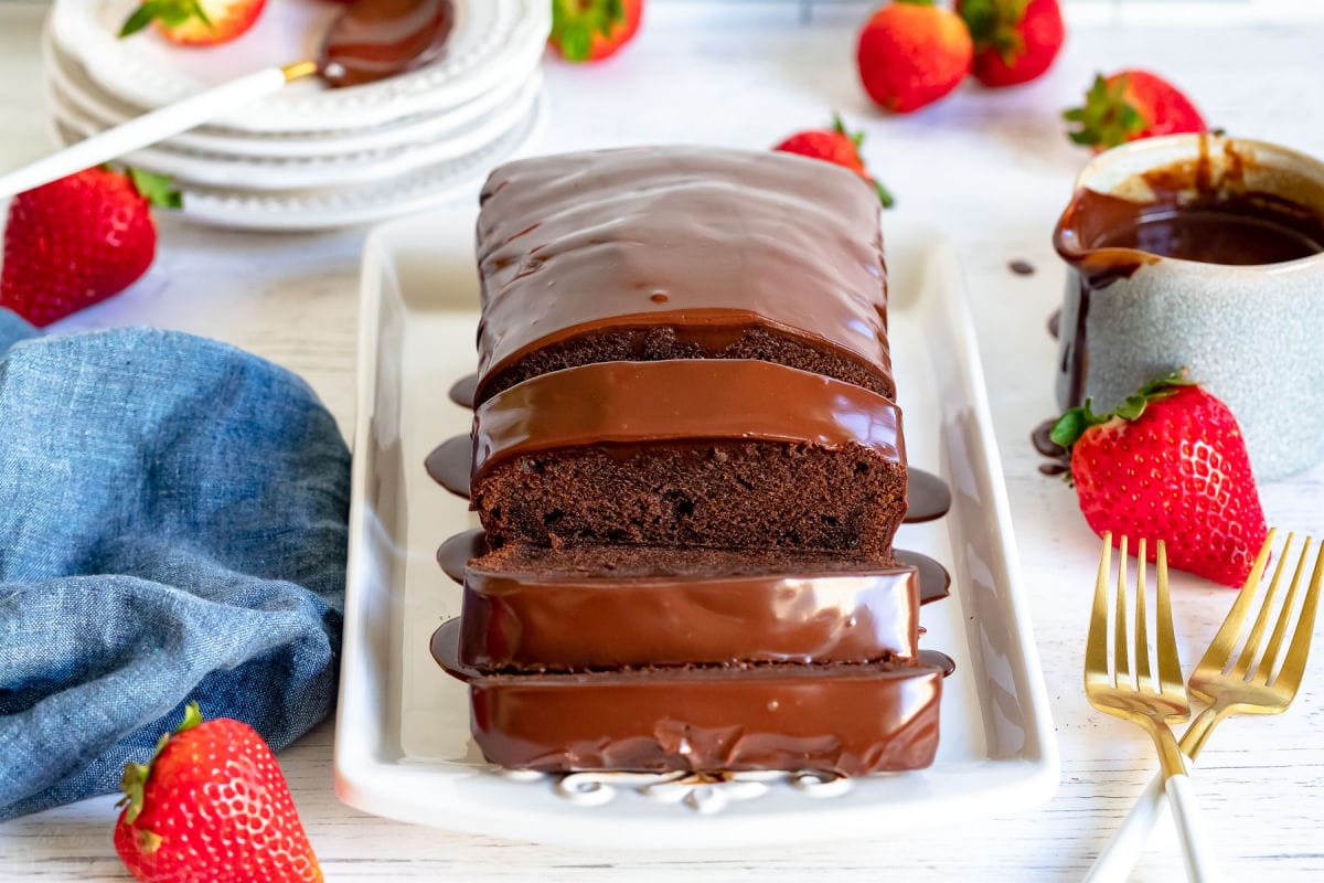 chocolate pound cake loaf with three slices cut on white tray with gold forks next to it and strawberries scattered about