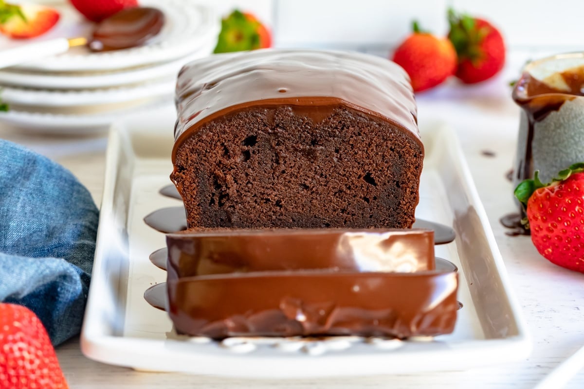 chocolate pound cake loaf with three slices cut on white platter with gold forks next to it and strawberries scattered about and blue napkin to the left