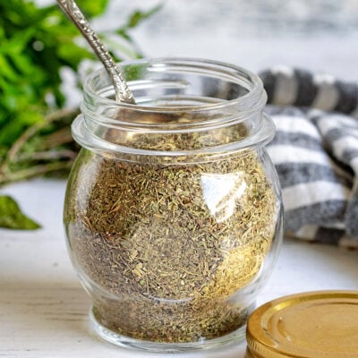 italian seasoning in a small glass jar with a spoon in the jar.