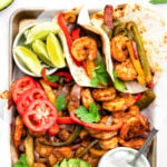 shrimp fajitas on a sheet pan with all the fixings including avocado lime wedges tomatoes and sour cream.