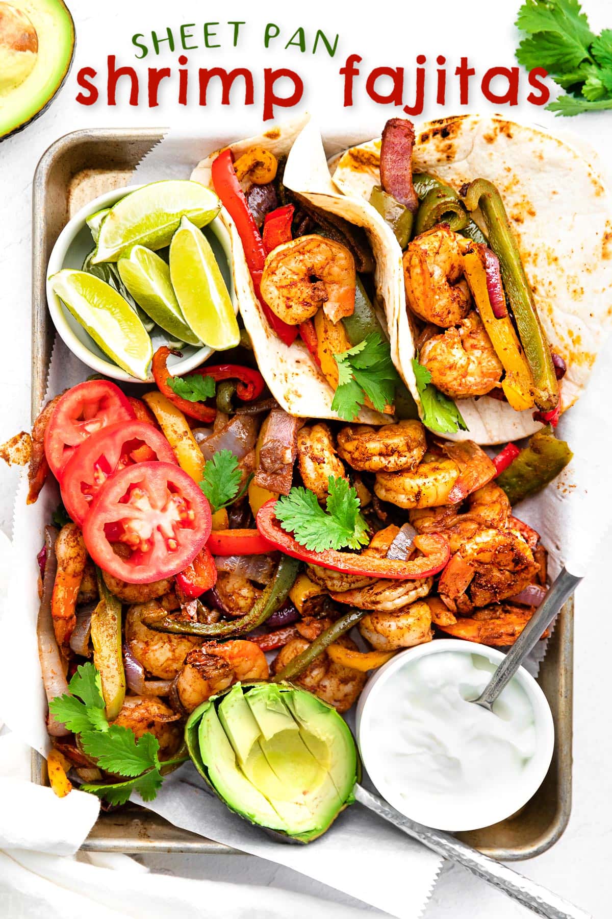 shrimp fajitas on a sheet pan with all the fixings including avocado lime wedges and sour cream.