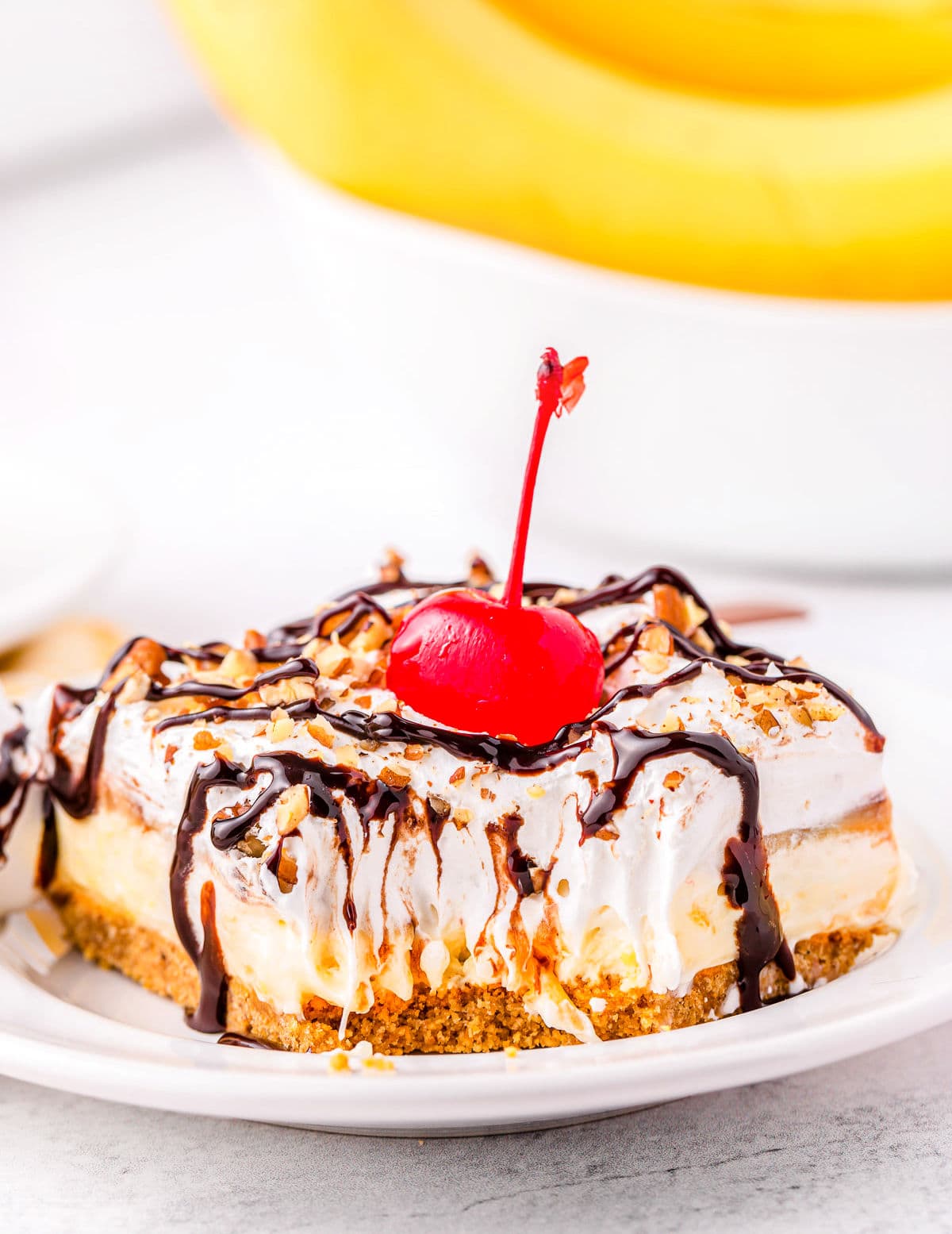 banana split dessert on white plate topped with chocolate sauce, nuts and a cherry.