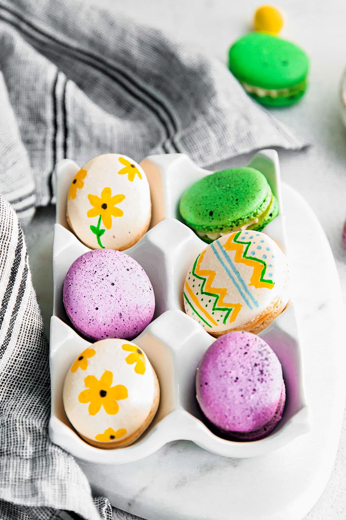 6 macarons decorated like eater eggs in white ceramic egg crate.