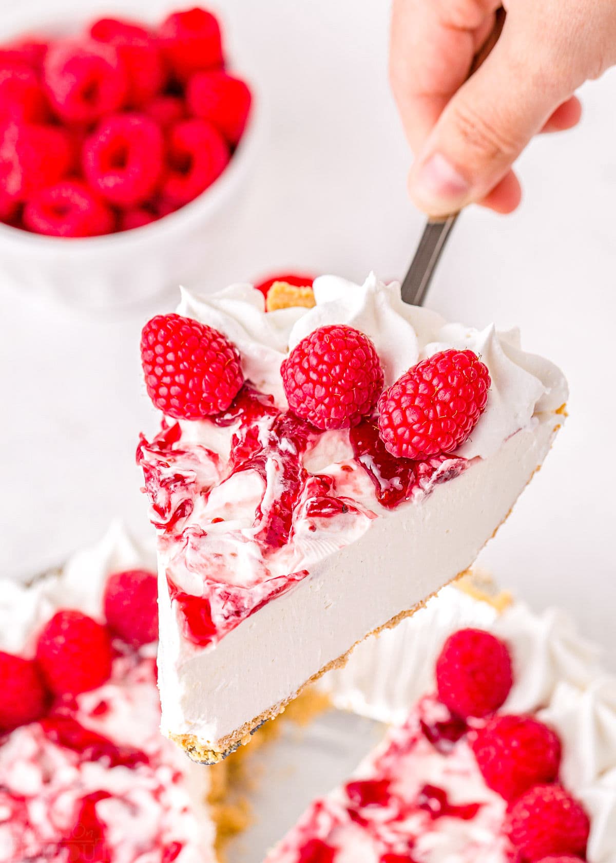 slice of no bake cheesecake with raspberry topping held by a pie server above cake.