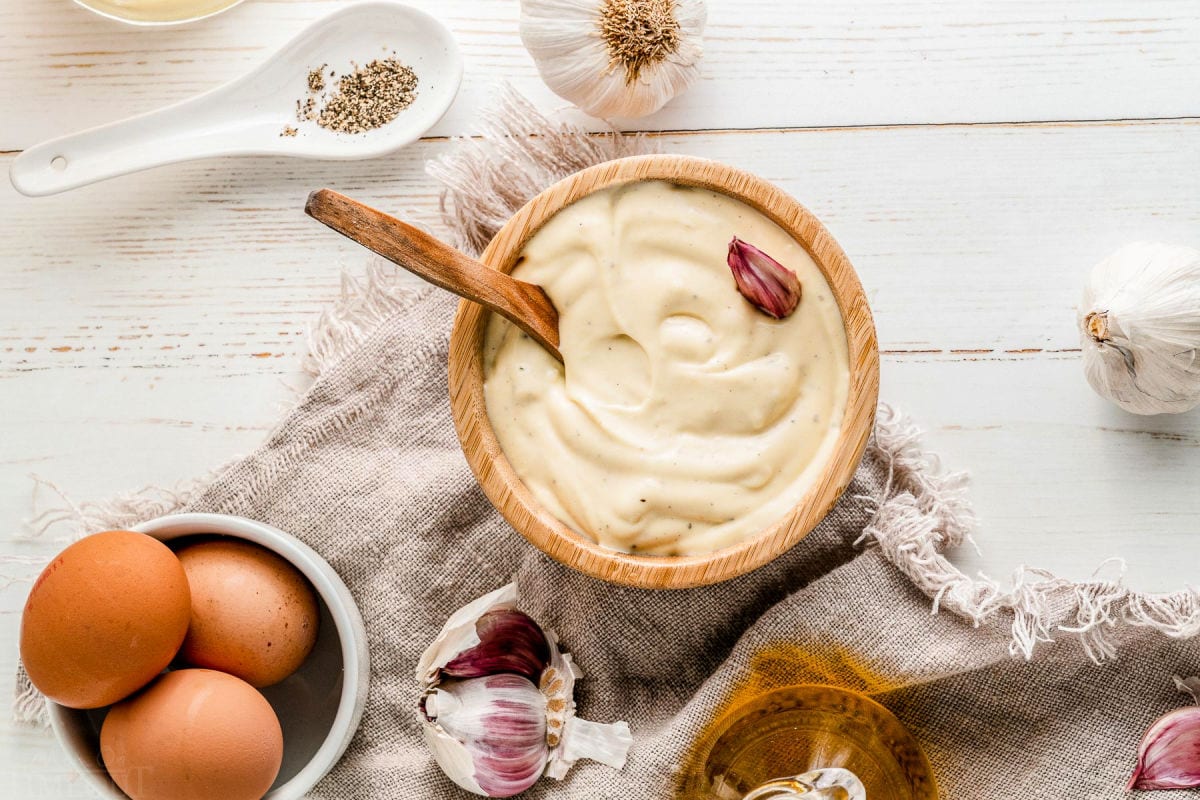 top down look at aioli recipe in small wooden bowl with wooden spoon in it. Aioli ingredients scattered around the outside of the bowl.