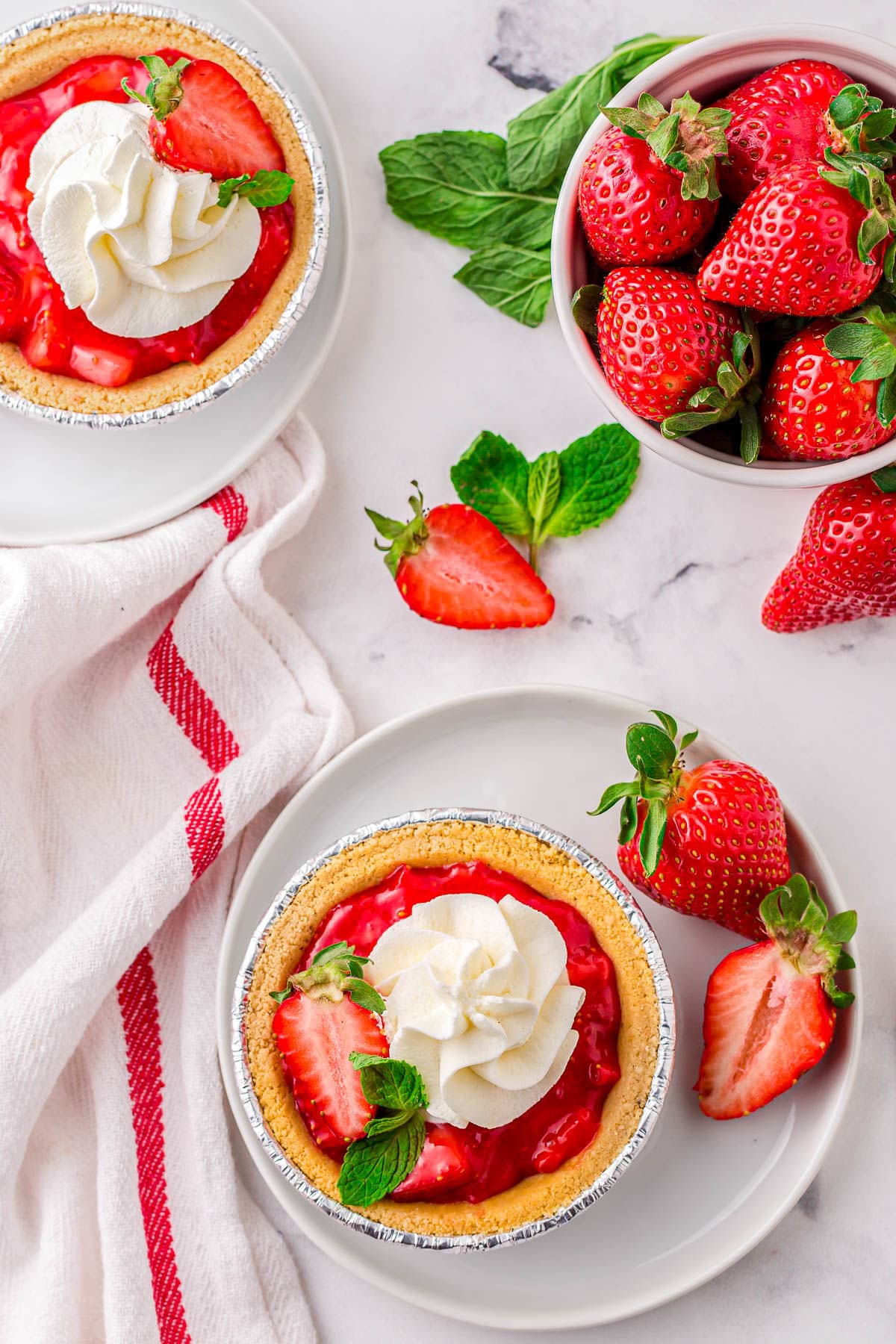 top down view of two mini strawberry pies on white plates with fresh strawberries scattered about.