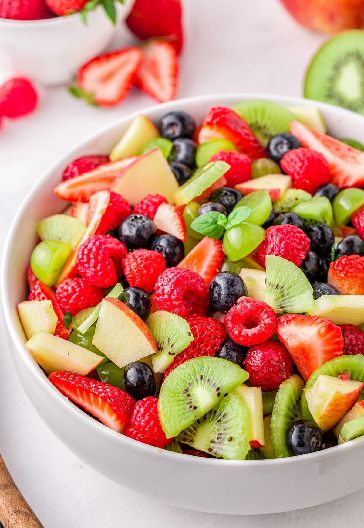 close up look at summer fruit salad in large white serving bowl. Salad has bright red strawberries, raspberries, green kiwis and grapes and blueberries.