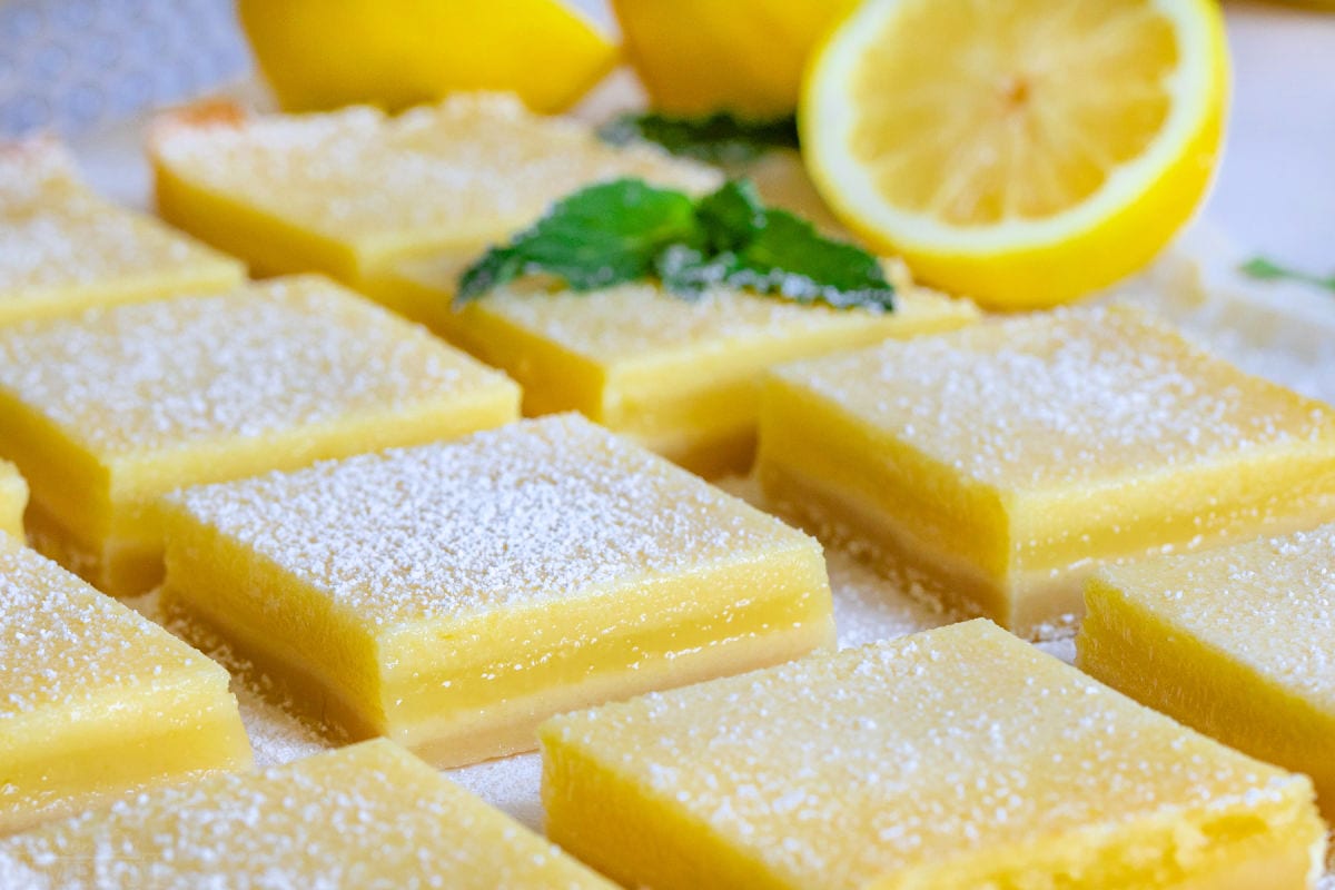 entire recipe of lemon bars cut up on white parchment with fresh lemons in background.