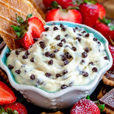 cannoli dip with chocolate chips sprinkled on top in a small white bowl with blue interior surrounded by cookies and strawberries ready to be enjoyed.
