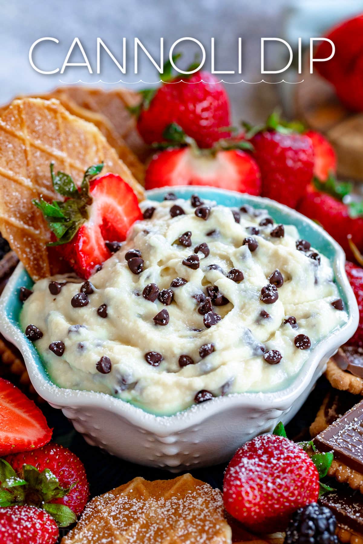cannoli dip in small white and blue bowl with strawberries and pizzelle cookies surrounding the bowl. title overlay at top of image.