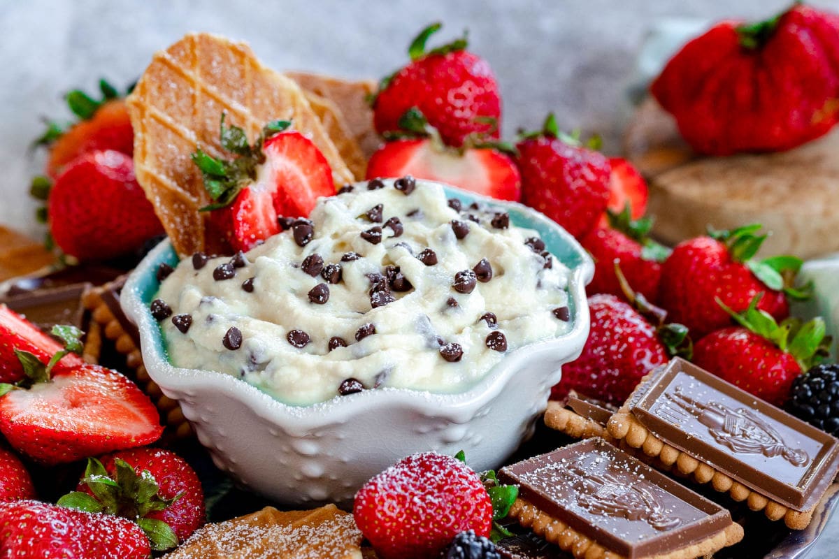 cannoli dip with chocolate chips in a small white bowl with blue interior surrounded by cookies and strawberries ready to be enjoyed.