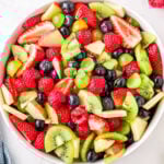 fruit salad in white bowl ready to serve with title overlay at top of image. fruit salad has kiwis, strawberries grapes and more.
