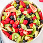 fruit salad in white bowl ready to serve with title overlay at top of image. fruit salad has kiwis, strawberries grapes and more.