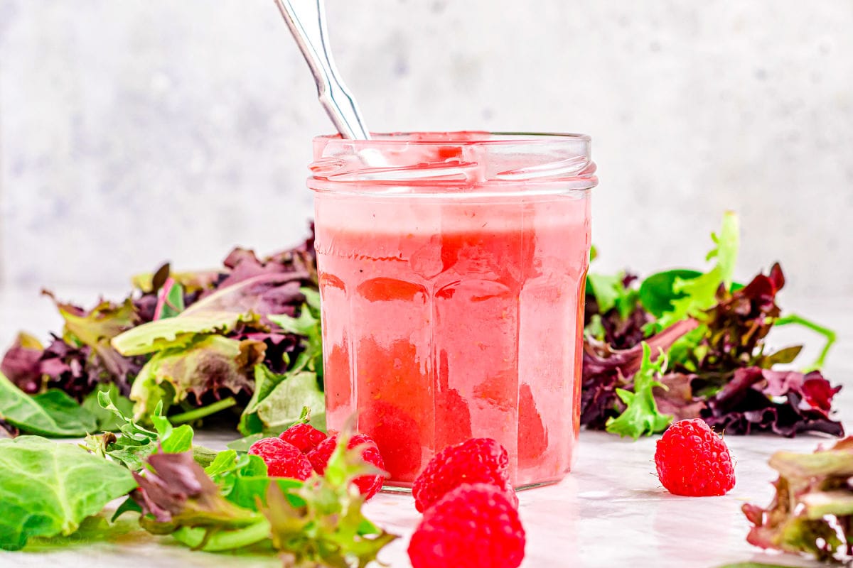 jar of raspberry vinaigrette sitting on white surface amongst greens and raspberries.